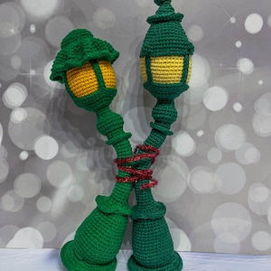 May include: Two green crocheted lampposts with yellow light fixtures are tied together with red ribbon. The lampposts are standing on a white surface.