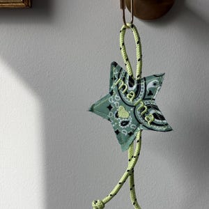 以下が含まれることがあります： 「mama」の刺繍が入った星型のキーホルダー。キーホルダーは緑色のバンダナ柄の生地とネオングリーンのロープでできています。キーホルダーは金色の金属製のバーから吊り下げられています。