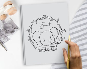 Puede incluir: Un libro gris claro con un dibujo de línea negra de un bebé elefante centrado dentro de una corona de hojas. El nombre Emmett está escrito en letra cursiva negra sobre el elefante. El libro está colocado junto a un bolígrafo dorado.