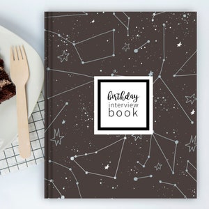 May include: A hardcover book with a dark gray cover featuring a white celestial constellation pattern and a central white square label with the black text birthday interview book. The book is positioned next to a plate with chocolate cake.