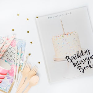 May include: A birthday-themed arrangement with a cake on a book titled "Birthday Interview Book." Pink paper cups, wooden cutlery, and floral stationery are arranged with gold star confetti. The cake has sprinkles and a lit candle.