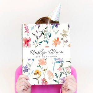 May include: A hardback birthday book with a white background featuring a colourful watercolour floral pattern. A central white horizontal band displays the text Kinsley Olivia BIRTHDAY BOOK in black script and sans-serif lettering.