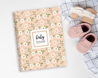 May include: A hardcover baby book with a peach-colored cover featuring a repeating pattern of white flowers and green leaves. A central white square with a peach border contains the black text baby book. Beside it are pink leather moccasins.