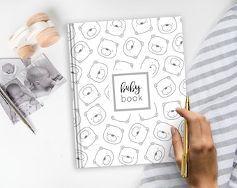 May include: A white hardcover baby book featuring a repeating black line art pattern of bear faces. A central gray square frame contains the black text baby book. The book is accompanied by a wooden toy, small black and white photos, and a gold pen.