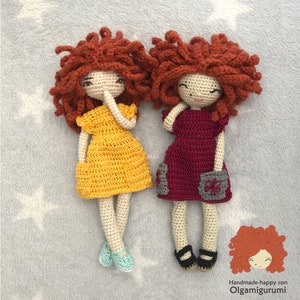 Puede incluir: Dos muñecas de crochet con pelo rojo rizado que llevan vestidos. Una muñeca lleva un vestido amarillo y la otra lleva un vestido burdeos. Ambas muñecas llevan zapatos.