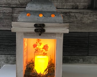 May include: White lantern with a metal top and a glass window. The window shows a lit candle, moss, and a small jar with flowers.