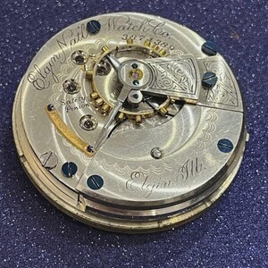 Op de afbeelding: Close-up van een uurwerk van een Elgin Nat'l Watch Co. zakhorloge. Het zilverkleurige mechanisme heeft ingewikkelde tandwielen, schroeven en gegraveerde details. Tekst bevat "Elgin Nat'l Watch Co.", "Safety Pinion" en "Elgin Ill."