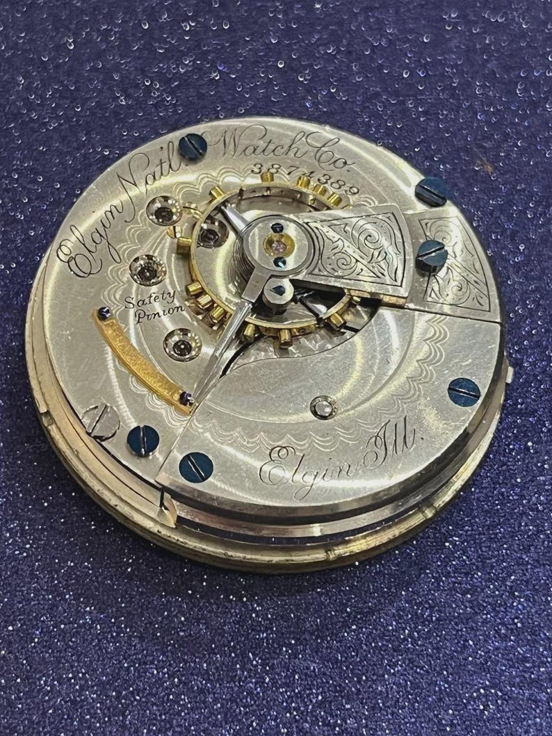 Op de afbeelding: Close-up van een uurwerk van een Elgin Nat'l Watch Co. zakhorloge. Het zilverkleurige mechanisme heeft ingewikkelde tandwielen, schroeven en gegraveerde details. Tekst bevat "Elgin Nat'l Watch Co.", "Safety Pinion" en "Elgin Ill."