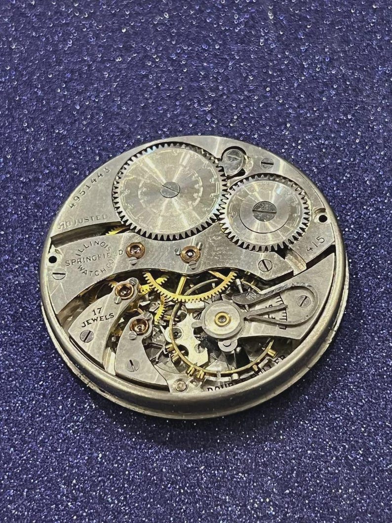 Puede incluir: Primer plano de un movimiento de reloj Illinois Springfield vintage. El mecanismo de esfera abierta presenta intrincados engranajes, joyas y el texto "17 Jewels". El reloj est&aacute; marcado como "Adjusted" y tiene el n&uacute;mero de serie 4951443.