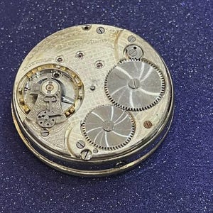 May include: Close-up of a vintage pocket watch mechanism. The silver-toned gears and intricate inner workings are visible, with the words "EAGLE LIMITED" and "15 JEWELS" etched on the metal. The watch is set against a dark blue background.