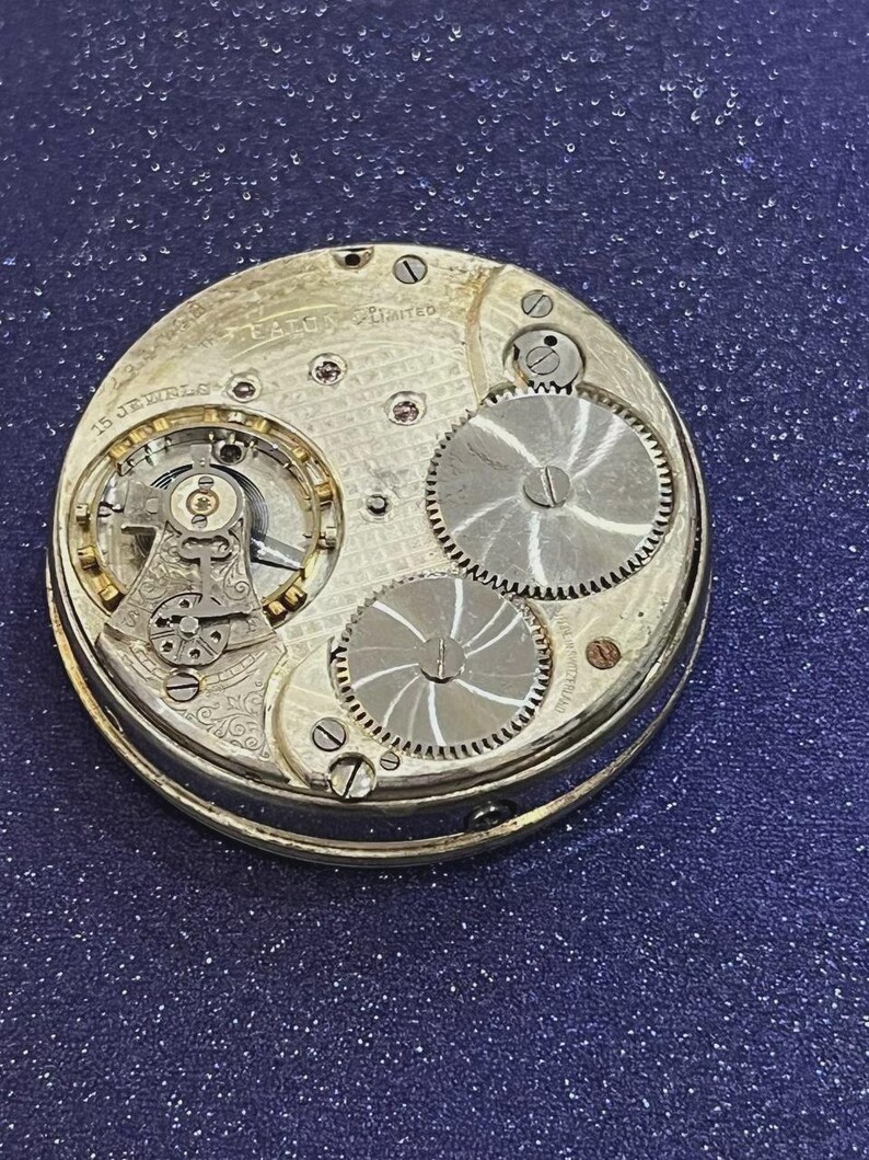 May include: Close-up of a vintage pocket watch mechanism. The silver-toned gears and intricate inner workings are visible, with the words "EAGLE LIMITED" and "15 JEWELS" etched on the metal. The watch is set against a dark blue background.