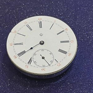May include: A vintage pocket watch face with a white dial, black Roman numerals, and a smaller seconds dial. The watch has black hands and a silver-colored metal casing. The numbers are marked in red at the 5, 10, 15, 20, 25, 30, 35, 40, 45, 50, 55, and 60-minute marks.