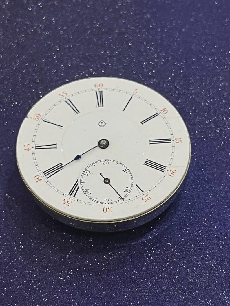 May include: A vintage pocket watch face with a white dial, black Roman numerals, and a smaller seconds dial. The watch has black hands and a silver-colored metal casing. The numbers are marked in red at the 5, 10, 15, 20, 25, 30, 35, 40, 45, 50, 55, and 60-minute marks.