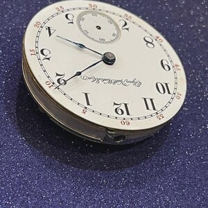 Op de afbeelding: Een vintage Elgin National Watch Co. zakhorlogekast. De witte wijzerplaat heeft zwarte Romeinse cijfers, een kleinere secondenwijzer en zwarte en blauwe wijzers. De horlogekast is zilverkleurig.