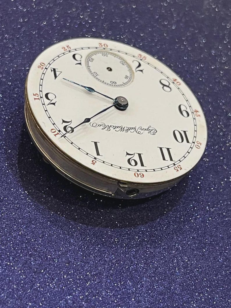 Op de afbeelding: Een vintage Elgin National Watch Co. zakhorlogekast. De witte wijzerplaat heeft zwarte Romeinse cijfers, een kleinere secondenwijzer en zwarte en blauwe wijzers. De horlogekast is zilverkleurig.