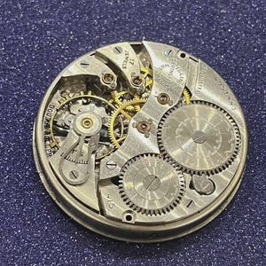 Puede incluir: Primer plano de un movimiento de reloj antiguo con intrincados engranajes y mecanismos. Los componentes plateados incluyen las palabras "17 Jewels" y "Springfield Watch Co.". El movimiento del reloj se encuentra sobre un fondo azul oscuro brillante.