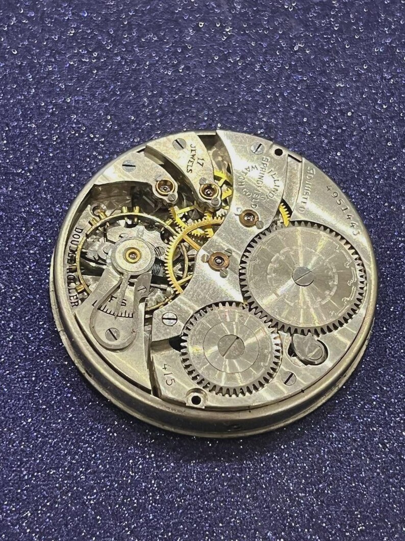 Puede incluir: Primer plano de un movimiento de reloj antiguo con intrincados engranajes y mecanismos. Los componentes plateados incluyen las palabras "17 Jewels" y "Springfield Watch Co.". El movimiento del reloj se encuentra sobre un fondo azul oscuro brillante.