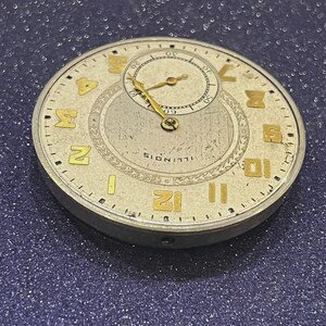 Puede incluir: Primer plano de la esfera de un reloj de bolsillo antiguo. La esfera es de color blanquecino con n&uacute;meros y agujas doradas. Una esfera m&aacute;s peque&ntilde;a est&aacute; en la parte superior. La palabra "ILLINOIS" est&aacute; impresa en la parte inferior.