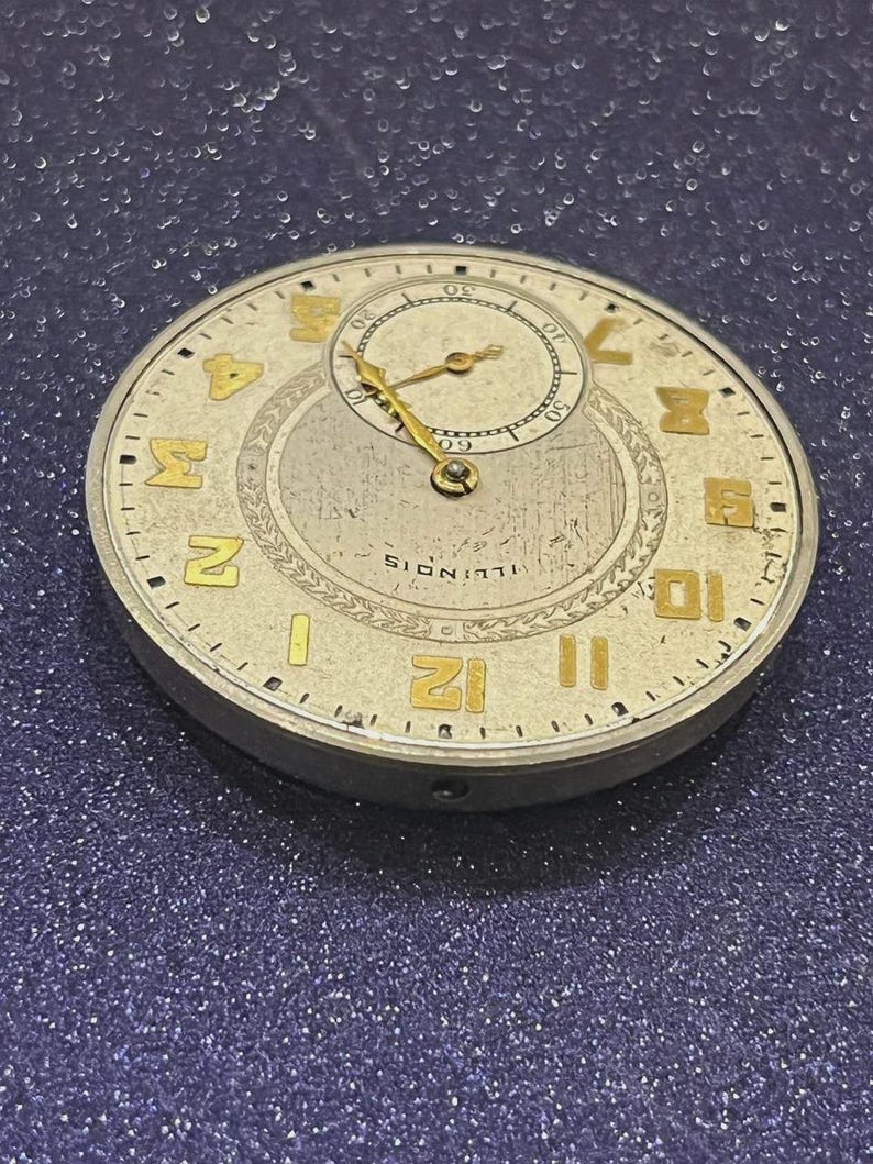 Puede incluir: Primer plano de la esfera de un reloj de bolsillo antiguo. La esfera es de color blanquecino con n&uacute;meros y agujas doradas. Una esfera m&aacute;s peque&ntilde;a est&aacute; en la parte superior. La palabra "ILLINOIS" est&aacute; impresa en la parte inferior.