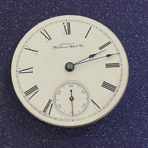 Może przedstawiać: Zbliżenie tarczy zabytkowego zegarka kieszonkowego. Tarcza jest biała z czarnymi rzymskimi cyframi i wskazówkami. Tekst "AMERICAN Waltham Watch Co." jest wydrukowany na czarno. Mniejsza tarcza pomocnicza wyświetla sekundy.