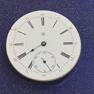 May include: A white pocket watch face with black Roman numerals and minute markers. A smaller subdial is at the bottom. The watch face has a small emblem near the center. The hands are black.