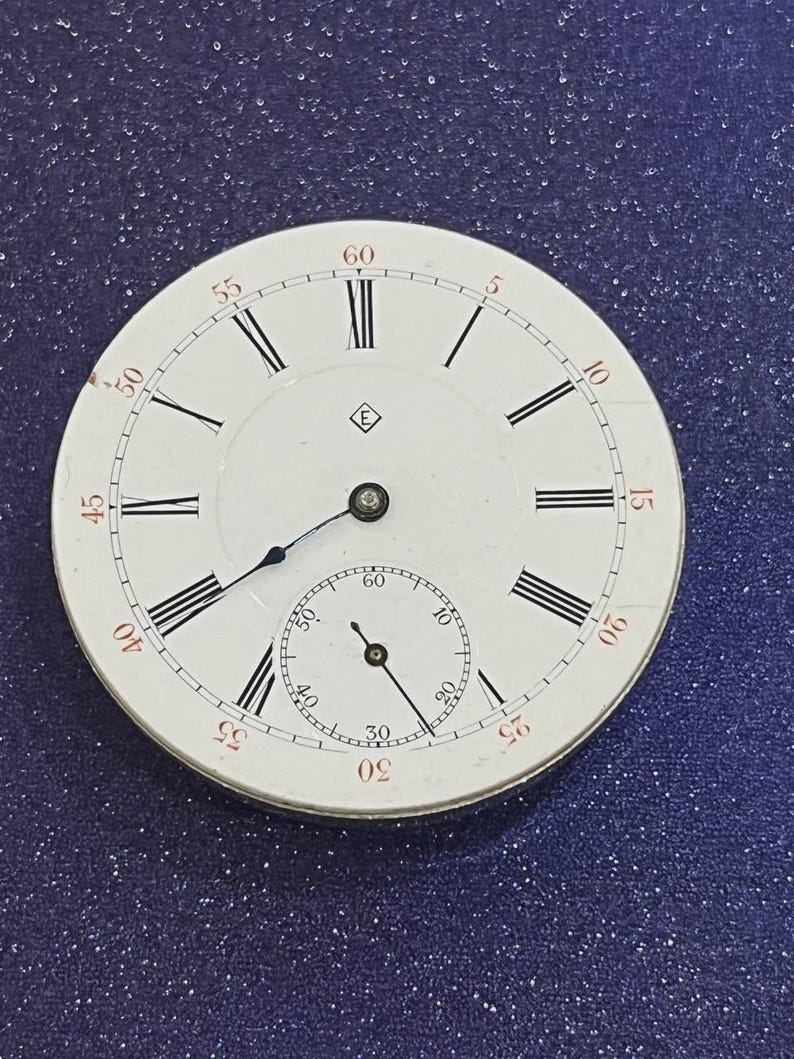 May include: A white pocket watch face with black Roman numerals and minute markers. A smaller subdial is at the bottom. The watch face has a small emblem near the center. The hands are black.