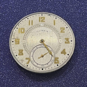 Puede incluir: Primer plano de una esfera de reloj vintage con n&uacute;meros y agujas doradas sobre fondo plateado. La palabra "ILLINOIS" es visible. Una esfera secundaria m&aacute;s peque&ntilde;a est&aacute; en la parte inferior. La esfera del reloj es redonda, aprox. 4 cm de di&aacute;metro.