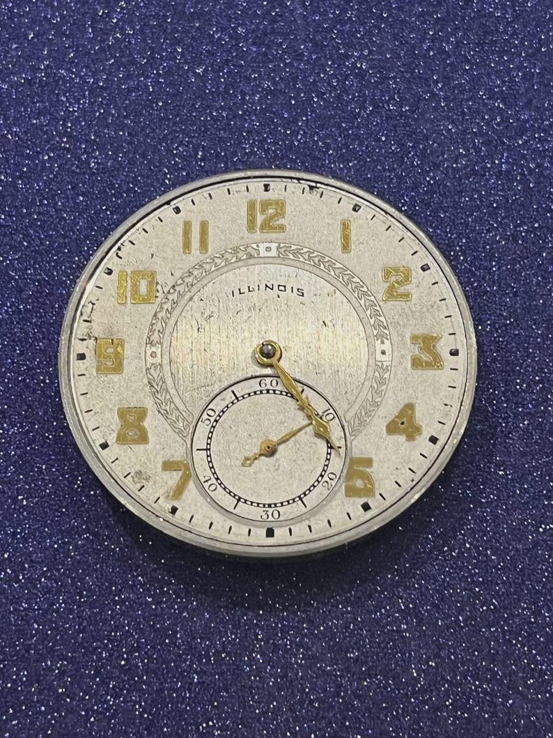 Puede incluir: Primer plano de una esfera de reloj vintage con n&uacute;meros y agujas doradas sobre fondo plateado. La palabra "ILLINOIS" es visible. Una esfera secundaria m&aacute;s peque&ntilde;a est&aacute; en la parte inferior. La esfera del reloj es redonda, aprox. 4 cm de di&aacute;metro.
