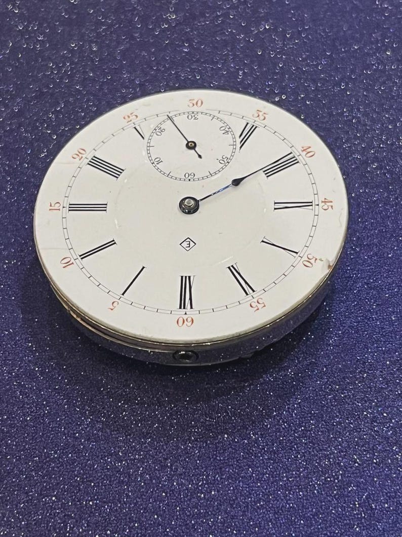 May include: A vintage pocket watch with a white face, black Roman numerals, and red minute markers. The watch has a small subdial and a silver-colored metal casing. The hands are black and blue.