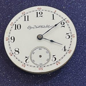 Op de afbeelding: Een vintage horlogekast met een witte achtergrond, zwarte cijfers en rode accenten. De horlogekast heeft de tekst "Elgin Natl Watch Co" en een kleinere subwijzerplaat. De wijzers zijn zwart.