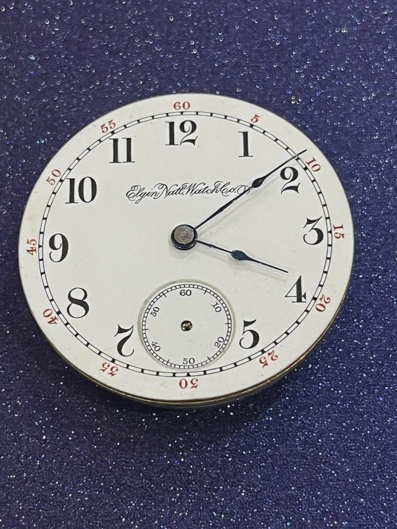 Op de afbeelding: Een vintage horlogekast met een witte achtergrond, zwarte cijfers en rode accenten. De horlogekast heeft de tekst "Elgin Natl Watch Co" en een kleinere subwijzerplaat. De wijzers zijn zwart.