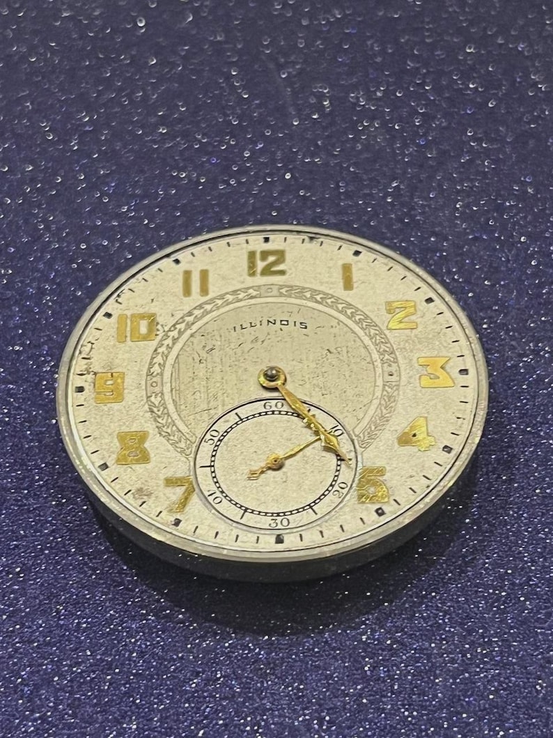 Puede incluir: Una esfera de reloj vintage con n&uacute;meros y manecillas doradas sobre un fondo color crema. La palabra "ILLINOIS" es visible en el centro. La esfera del reloj tiene una esfera secundaria m&aacute;s peque&ntilde;a en la parte inferior.