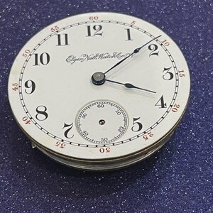 Op de afbeelding: Een vintage Elgin Natl Watch Co. zakhorlogeklok. De witte wijzerplaat heeft zwarte Arabische cijfers, een kleine secondenwijzer en zwarte wijzers. Rode minutenmarkeringen omzomen de rand. Het horloge staat tegen een donkerblauwe achtergrond.