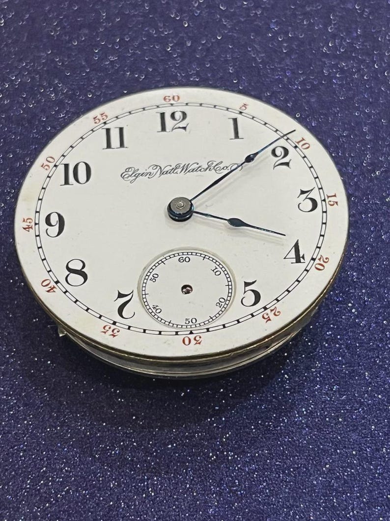 Op de afbeelding: Een vintage Elgin Natl Watch Co. zakhorlogeklok. De witte wijzerplaat heeft zwarte Arabische cijfers, een kleine secondenwijzer en zwarte wijzers. Rode minutenmarkeringen omzomen de rand. Het horloge staat tegen een donkerblauwe achtergrond.