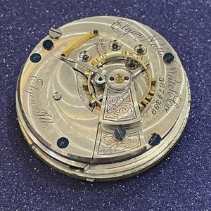 Op de afbeelding: Close-up van een antiek Elgin National Watch Co. zakhorlogemechanisme. Het zilverkleurige mechanisme heeft ingewikkelde gravures, zichtbare tandwielen en de tekst "Elgin Nat'l Watch Co." en "3874388". Het horloge heeft een goudkleurige balansveer en schroeven.