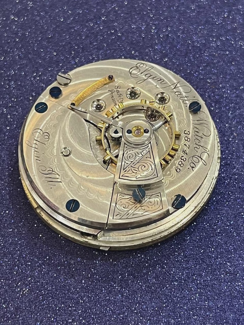 Op de afbeelding: Close-up van een antiek Elgin National Watch Co. zakhorlogemechanisme. Het zilverkleurige mechanisme heeft ingewikkelde gravures, zichtbare tandwielen en de tekst "Elgin Nat'l Watch Co." en "3874388". Het horloge heeft een goudkleurige balansveer en schroeven.