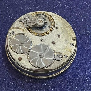 May include: Close-up of a vintage watch movement with intricate gears and components. The watch mechanism features a silver-colored metal casing with engraved details and visible jewels. The design includes two large, circular gears and smaller, detailed mechanisms.
