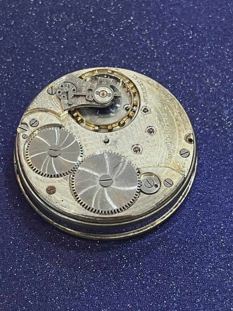 May include: Close-up of a vintage watch movement with intricate gears and components. The watch mechanism features a silver-colored metal casing with engraved details and visible jewels. The design includes two large, circular gears and smaller, detailed mechanisms.