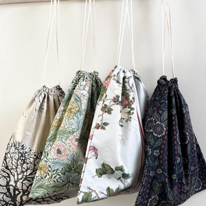 May include: Four fabric drawstring bags hanging from a wooden rack. The first bag is beige with a black tree silhouette. The second features a green, pink, and yellow floral pattern. The third is white with red and green floral motifs. The fourth is dark blue with a dense, multi-coloured botanical print.