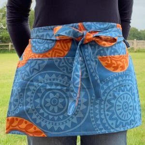 May include: A blue and orange patterned half apron with a bow tie. The apron features a repeating circular design in shades of blue and orange. The apron is tied at the waist and has two pockets.