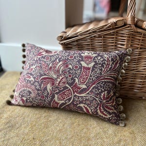 May include: A rectangular decorative pillow with a floral pattern in shades of purple, burgundy, and cream. The pillow has a pom-pom trim and rests on a yellow herringbone fabric. A wicker basket is in the background.