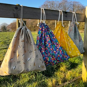 May include: Four drawstring bags hanging from a wooden fence. The first is beige with animal prints, the second features a colourful abstract pattern, the third is solid yellow with white sun icons, and the fourth displays a vintage world map print.