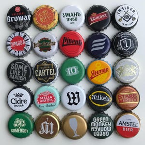 May include: A collection of 25 colorful bottle caps with various logos and text. The caps are round and metallic, featuring brands like Stella Artois, Amstel, and Green Monkey. Some caps have text in Cyrillic script.