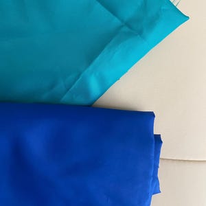 May include: Two folded pieces of fabric are displayed. The top piece is a vibrant turquoise blue, while the bottom piece is a deep, saturated royal blue. Both fabrics have a smooth, slightly reflective surface texture and are neatly layered.