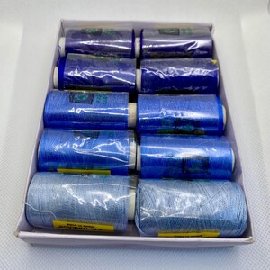 May include: A white cardboard box containing ten spools of sewing thread arranged in five rows of two. The spools feature a gradient of blue shades, ranging from deep navy at the top to a light, pale blue at the bottom, all wrapped in clear plastic.