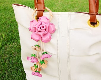 May include: A white tote bag with brown leather handles and a pink leather rose charm. The rose charm has a green stem and leaves.