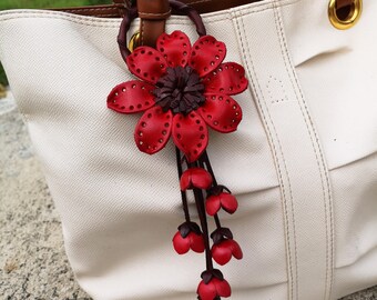Könnte beinhalten: Ein roter Lederblumenanhänger mit brauner Mitte und einer Kaskade kleinerer roter Lederblumen, die von unten hängen. Der Anhänger ist an einer weißen Tragetasche mit braunen Ledergriffen befestigt.