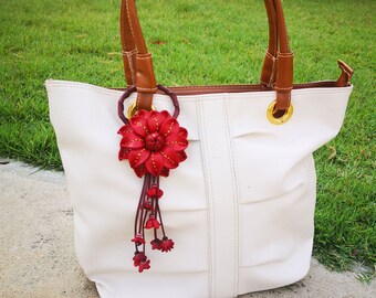 Könnte beinhalten: Eine weiße Tragetasche mit braunen Ledergriffen und einem roten Lederblumen-Schlüsselanhänger. Die Tasche hat einen goldenen Reißverschluss und goldene Ösen.