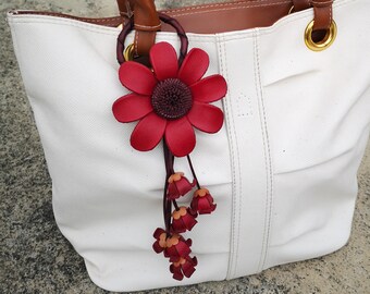 Könnte beinhalten: Eine weiße Tragetasche mit braunen Ledergriffen und einem roten Lederblumenanhänger. Der Blumenanhänger hat eine burgunderrote Mitte und einen langen Stiel mit kleineren roten Blüten.