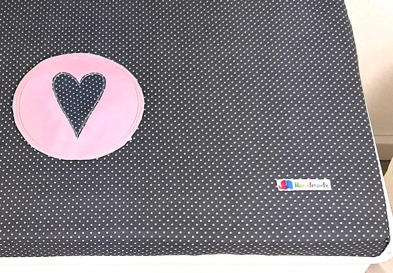 May include: A black changing pad with white polka dots and a pink circle with a black heart in the center. The changing pad has a white label that says "Handmade".